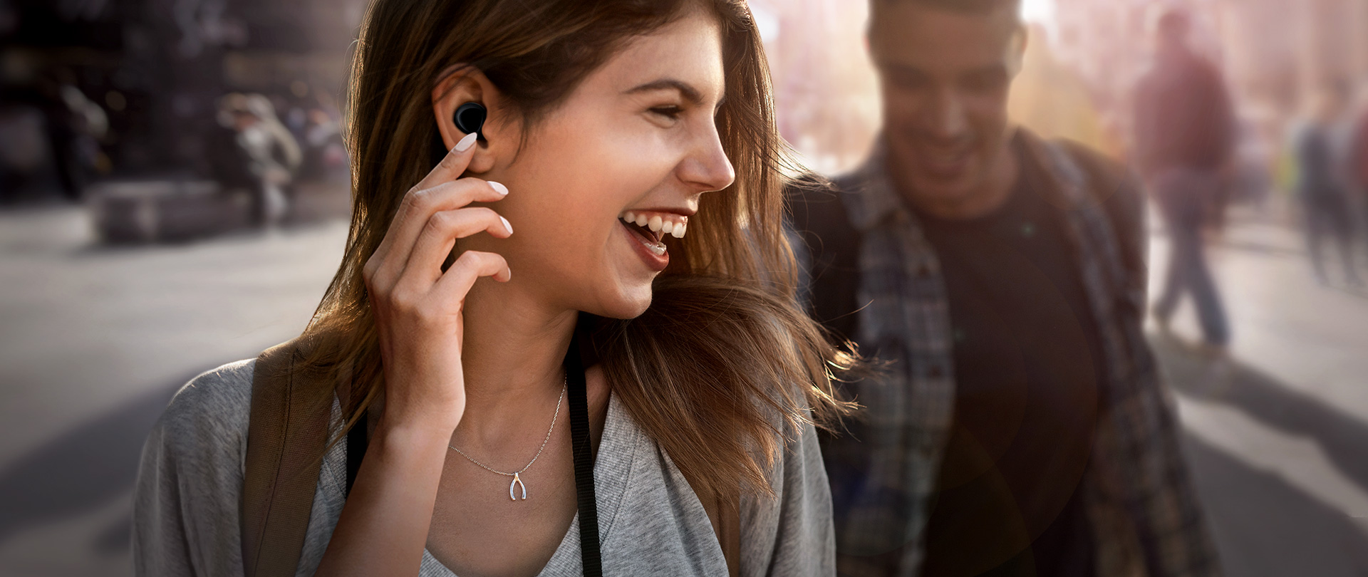 A woman wearing Galaxy Buds in the background of a crowded city is touching the product in her ear with her fingers and a man is standing behind her.