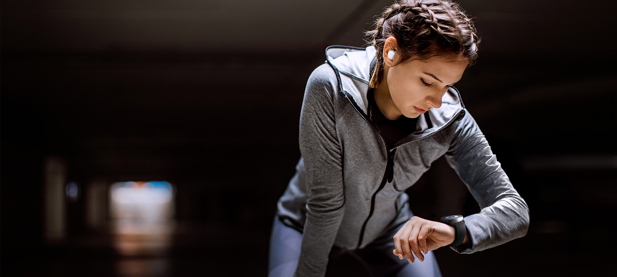 A woman wearing a white Galaxy Buds is holding a hand on her knee and watching a black Galaxy Watch Active on her wrist.