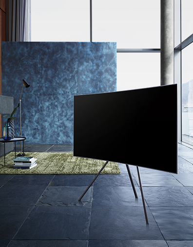 파란색감의 인테리어로 구성이 된 실내중앙에 스튜디오 스탠드가 장착된 QLED TV가 우측으로 비스듬하게 가운데에 세워져 있습니다.