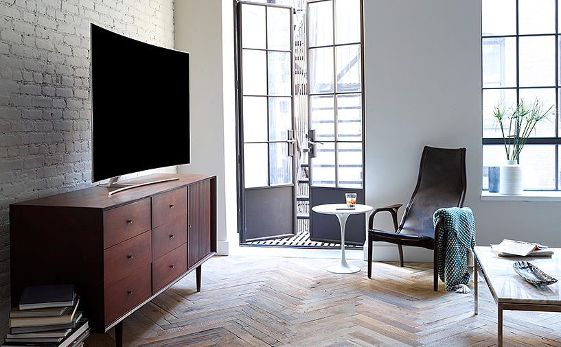 선반위에 올라가져 있는 QLED TV의 측면이 보여지고 있으며 제품의 앞에는 쇼파 및 테이블이 보여집니다.