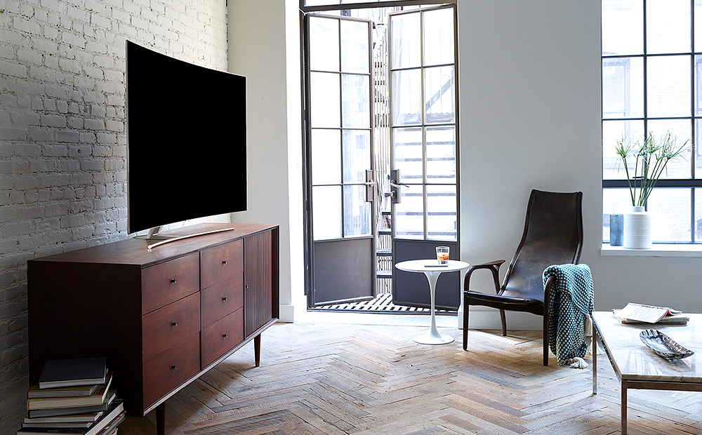 선반위에 올라가져 있는 QLED TV의 측면이 보여지고 있으며 제품의 앞에는 쇼파 및 테이블이 보여집니다.