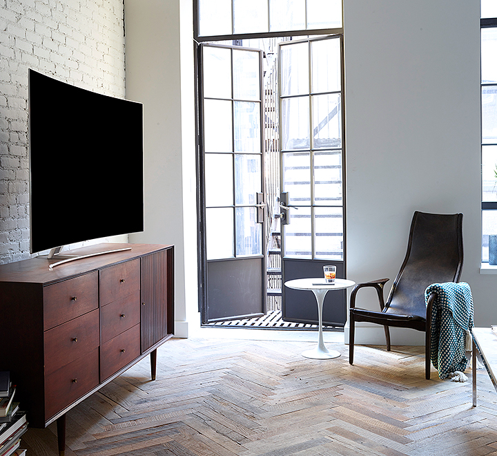 선반위에 올라가져 있는 QLED TV의 측면이 보여지고 있으며 제품의 앞에는 쇼파 및 테이블이 보여집니다.